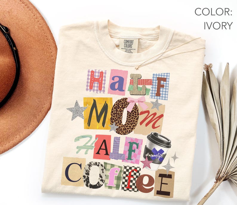 May include: Ivory-colored t-shirt with the words "Half Mom Half Coffee" in a collage style. The design includes a coffee cup graphic and star accents. The shirt is made of a soft material and is a Comfort Colors brand.