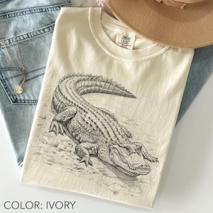 May include: Ivory-colored t-shirt featuring a detailed black and white illustration of an alligator. The alligator is depicted in a realistic style, lying down. The shirt is paired with blue jeans and a necklace. The text "COLOR: IVORY" is at the bottom.