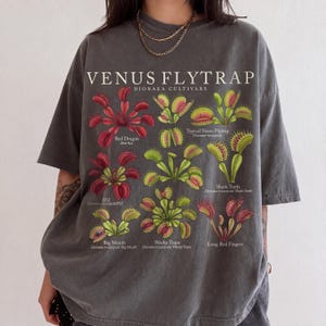 May include: A charcoal gray t-shirt featuring a botanical illustration of Venus flytrap cultivars. The graphic includes the text VENUS FLYTRAP DIONAEA CULTIVARS and labels for Red Dragon, Typical Venus Flytrap, Shark Teeth, B52, Big Mouth, Wacky Traps, and Long Red Fingers.