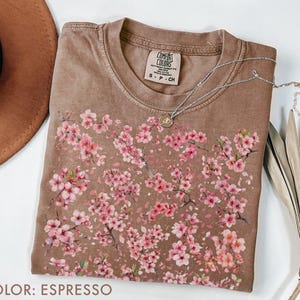 May include: Espresso-colored t-shirt with a crew neck, featuring a floral design of pink blossoms. The shirt has a Comfort Colors tag and a silver necklace with a pendant. A brown hat and dried palm leaf are also visible.