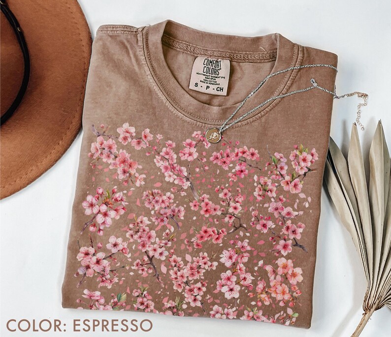 May include: Espresso-colored t-shirt with a crew neck, featuring a floral design of pink blossoms. The shirt has a Comfort Colors tag and a silver necklace with a pendant. A brown hat and dried palm leaf are also visible.