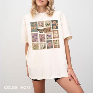 K&ouml;nnte beinhalten: Elfenbeinfarbenes T-Shirt mit einer Sammlung von Briefmarken im Vintage-Stil. Die Briefmarken zeigen Blumen- und Schmetterlingsdesigns in verschiedenen Farben. Das Shirt hat kurze &Auml;rmel und eine lockere Passform. Der Text "COLOR: IVORY" steht unten.