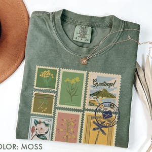 以下が含まれることがあります： モスグリーンのTシャツ。ヴィンテージ風の切手のコラージュが特徴で、花柄や風景のデザインが施されています。Tシャツはクルーネックで半袖です。小さなペンダントが付いたゴールドのネックレスがTシャツの上にあります。