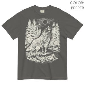 Puede incluir: Una camiseta de manga corta color pimienta con una ilustraci&oacute;n gr&aacute;fica blanca de un lobo aullando a la luna llena en un entorno forestal con pinos, monta&ntilde;as y un arroyo rocoso. El texto COLOR: PEPPER aparece en la esquina superior derecha.