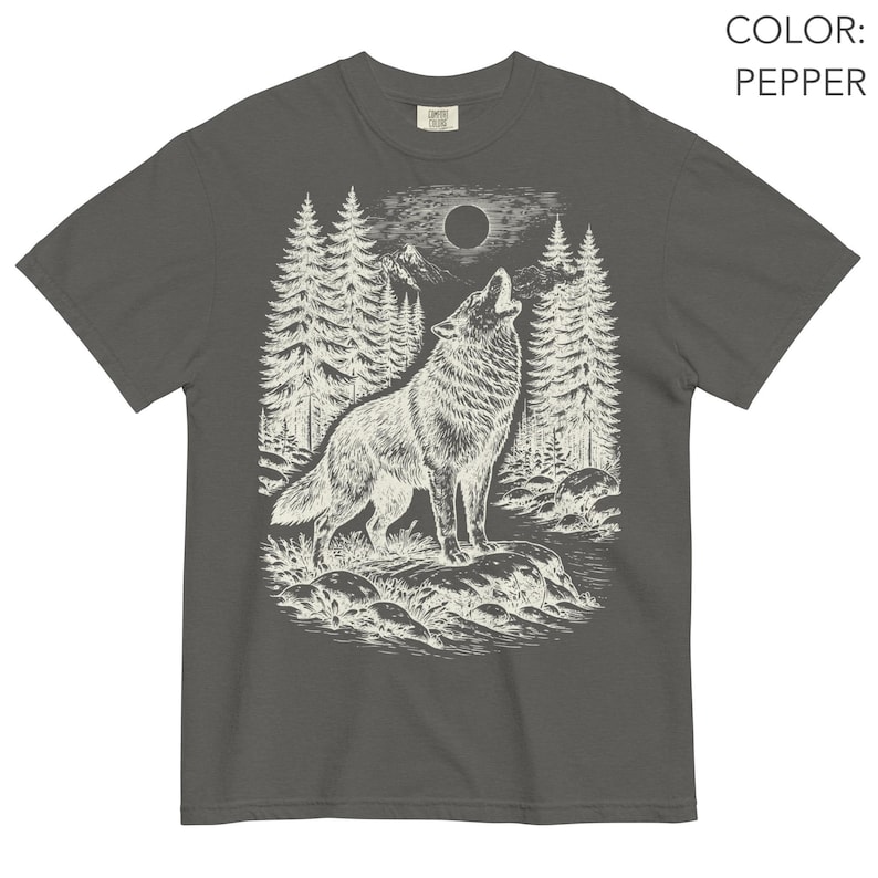 Puede incluir: Una camiseta de manga corta color pimienta con una ilustraci&oacute;n gr&aacute;fica blanca de un lobo aullando a la luna llena en un entorno forestal con pinos, monta&ntilde;as y un arroyo rocoso. El texto COLOR: PEPPER aparece en la esquina superior derecha.