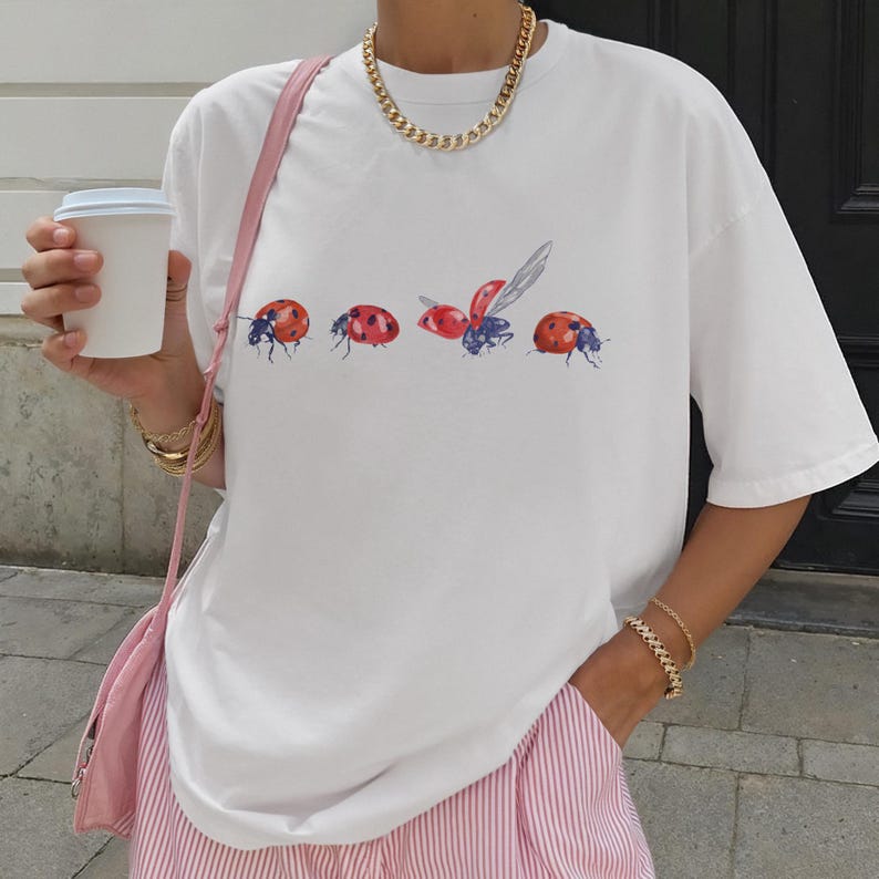 May include: White t-shirt with a series of red ladybug illustrations with black and blue details. The ladybugs are in various stages of flight. The shirt has short sleeves and a relaxed fit. The person is wearing a gold chain necklace and bracelets.