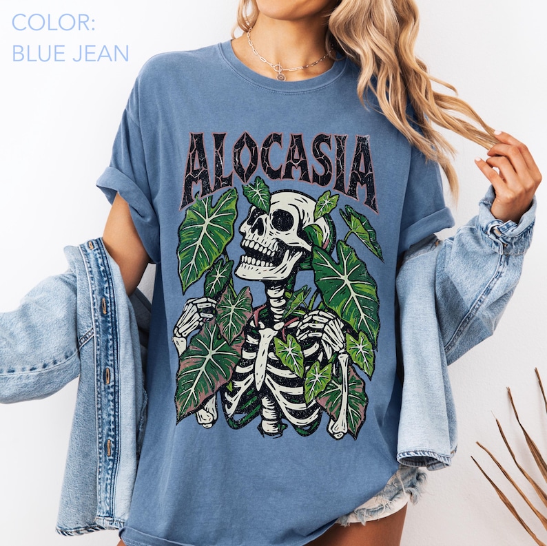 K&ouml;nnte beinhalten: Blaues Jeans-T-Shirt mit dem Wort "ALOCASIA" in brauner, verwaschener Schrift. Die Grafik zeigt ein Skelett, das von gr&uuml;nen Bl&auml;ttern umgeben ist. Das Shirt hat kurze &Auml;rmel und eine lockere Passform.