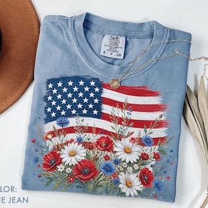 May include: Blue jean coloured t-shirt featuring a distressed American flag design with red, white, and blue flowers. The shirt has a crew neck and short sleeves. A gold necklace is draped across the shirt.