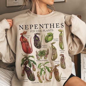 Op de afbeelding: Een beige sweater met ronde hals voorzien van een botanische illustratie van diverse Nepenthes bekerplanten. De tekst luidt "NEPENTHES PITCHER PLANTS" met labels voor de soorten Nepenthes rajah, rafflesiana, ampullaria, glacilis, albomarginata, truncata, alata en bicalcarata.