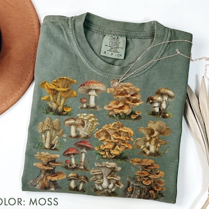 Pu&ograve; includere: T-shirt verde muschio con un'illustrazione dettagliata di vari funghi. Il design include una variet&agrave; di tipi di funghi in tonalit&agrave; di giallo, marrone e rosso. La maglietta ha un girocollo classico e maniche corte. Il testo "COLOR: MOSS" &egrave; in basso.