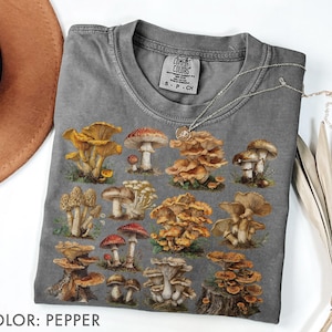 Pu&ograve; includere: T-shirt color pepe con un'illustrazione dettagliata di vari funghi nelle tonalit&agrave; del marrone, giallo e rosso. La maglietta ha un girocollo classico e maniche corte. L'etichetta recita "Comfort Colors". Una collana d'argento &egrave; drappeggiata sulla maglietta.