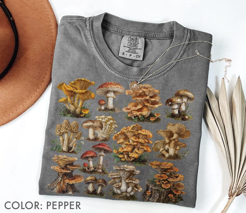 Pu&ograve; includere: T-shirt color pepe con un'illustrazione dettagliata di vari funghi nelle tonalit&agrave; del marrone, giallo e rosso. La maglietta ha un girocollo classico e maniche corte. L'etichetta recita "Comfort Colors". Una collana d'argento &egrave; drappeggiata sulla maglietta.