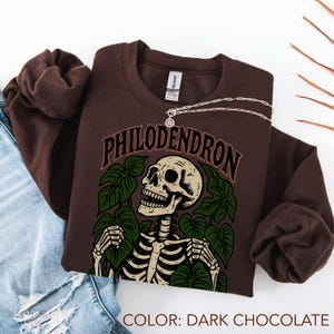 May include: Dark chocolate-colored sweatshirt with a graphic of a skeleton surrounded by green leaves and the word "PHILODENDRON". A silver necklace with a pendant is draped across the sweatshirt. The color of the sweatshirt is dark chocolate.