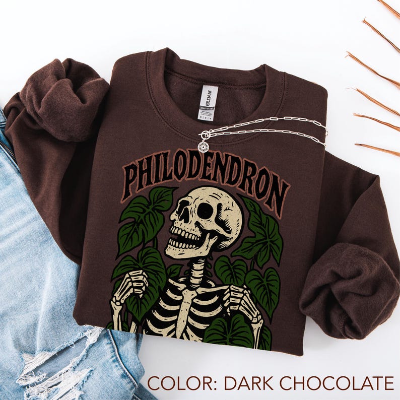 May include: Dark chocolate-colored sweatshirt with a graphic of a skeleton surrounded by green leaves and the word "PHILODENDRON". A silver necklace with a pendant is draped across the sweatshirt. The color of the sweatshirt is dark chocolate.