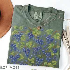 May include: A moss-colored t-shirt featuring a watercolor design of purple grapes on vines with green leaves. A silver necklace with a small pendant rests on the shirt. The text "COLOR: MOSS" is at the bottom.