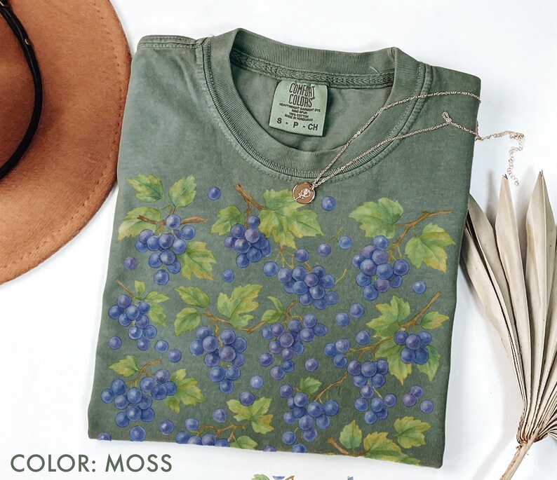 May include: A moss-colored t-shirt featuring a watercolor design of purple grapes on vines with green leaves. A silver necklace with a small pendant rests on the shirt. The text "COLOR: MOSS" is at the bottom.