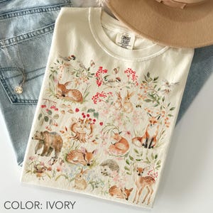 Peut inclure: Un t-shirt ivoire orné d'une impression style aquarelle représentant divers animaux des bois, dont un ours, un renard, des cerfs, des lapins et un hérisson, entourés d'éléments botaniques comme des baies rouges, des feuilles vertes et des fleurs.