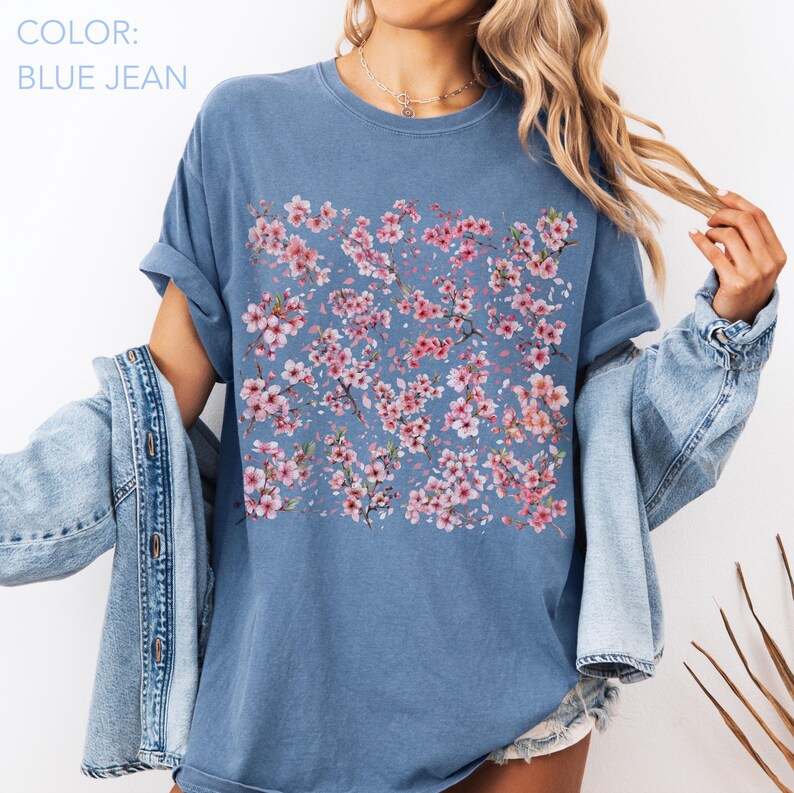 May include: Blue jean colored t-shirt with a floral pattern of pink cherry blossoms. The shirt has a crew neck and short sleeves. The text "COLOR: BLUE JEAN" is in the upper left corner.
