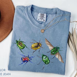 以下が含まれることがあります： クルーネックのブルーデニムカラーTシャツ。カラフルなカブトムシの水彩デザインが特徴です。ペンダント付きのゴールドネックレスがシャツの上にあります。シャツのタグには「Comfort Colors」と書かれています。