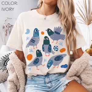 May include: Ivory-colored t-shirt featuring a playful design of several cartoon pigeons in shades of blue, gray, and green. The birds are surrounded by orange circles, blue shapes, and small blue and green leaf-like elements. The shirt is a casual, comfortable style.