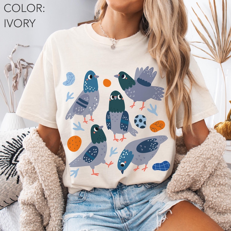 May include: Ivory-colored t-shirt featuring a playful design of several cartoon pigeons in shades of blue, gray, and green. The birds are surrounded by orange circles, blue shapes, and small blue and green leaf-like elements. The shirt is a casual, comfortable style.