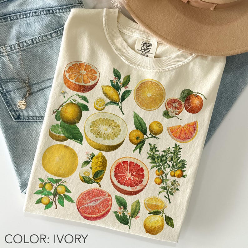 May include: An ivory t-shirt featuring a botanical print of various citrus fruits including oranges, grapefruits, and lemons. The graphic displays whole and sliced fruits with green leaves and small white blossoms. The shirt has a crew neckline.