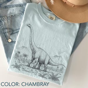 Pu&ograve; includere: T-shirt blu chambray con un'illustrazione in bianco e nero di un dinosauro Brachiosaurus. Il design include un paesaggio con palme e montagne. La maglietta &egrave; piegata con un cappello e jeans.