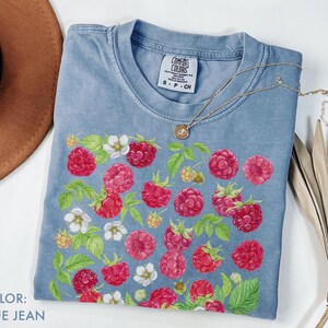 Op de afbeelding: Blauw spijkerbroekkleurig T-shirt met een frambozen- en bloemenontwerp. Het shirt heeft een ronde hals en korte mouwen. Een gouden ketting met een hanger is over het shirt gedrapeerd. De tekst "COLOR: BLUE JEAN" is linksonder zichtbaar.