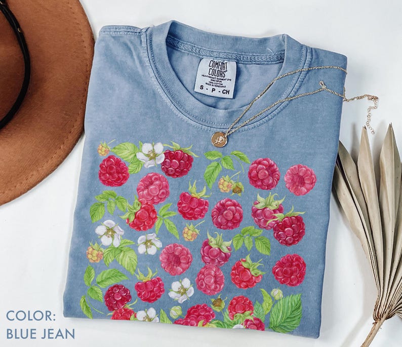 Op de afbeelding: Blauw spijkerbroekkleurig T-shirt met een frambozen- en bloemenontwerp. Het shirt heeft een ronde hals en korte mouwen. Een gouden ketting met een hanger is over het shirt gedrapeerd. De tekst "COLOR: BLUE JEAN" is linksonder zichtbaar.