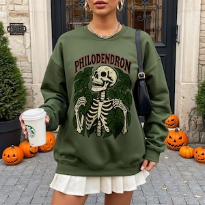 May include: Olive green sweatshirt featuring a skeleton graphic with the word "PHILODENDRON" above it. The skeleton is surrounded by green leaves. The sweatshirt is a long-sleeved, crew neck style.