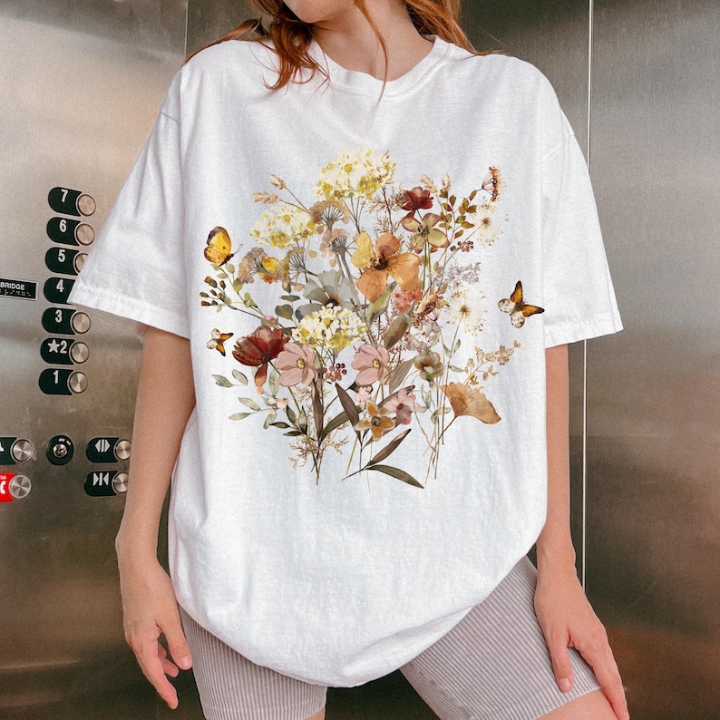 May include: White t-shirt featuring a floral bouquet design with butterflies. The bouquet includes flowers in shades of yellow, orange, and red. The butterflies add a whimsical touch to the design. The shirt is a casual, oversized fit.
