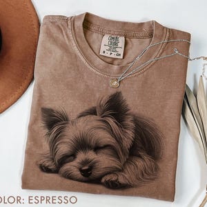 Puede incluir: Camiseta color espresso con una ilustraci&oacute;n detallada de un Yorkshire Terrier durmiendo. La camiseta tiene cuello redondo y una peque&ntilde;a etiqueta en el pecho. Un collar plateado con un peque&ntilde;o colgante descansa sobre la camiseta.