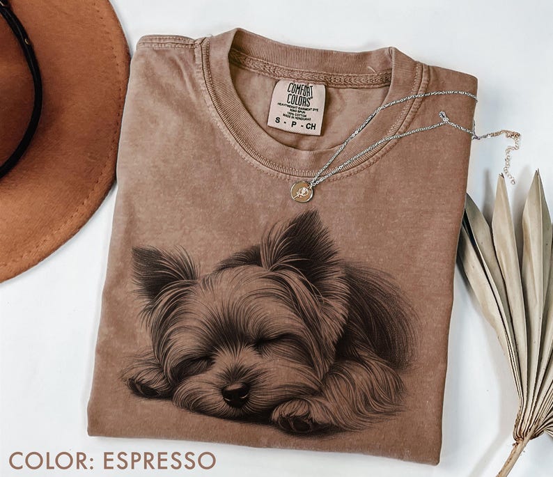 Puede incluir: Camiseta color espresso con una ilustraci&oacute;n detallada de un Yorkshire Terrier durmiendo. La camiseta tiene cuello redondo y una peque&ntilde;a etiqueta en el pecho. Un collar plateado con un peque&ntilde;o colgante descansa sobre la camiseta.