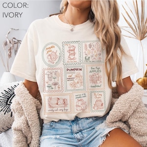 May include: Ivory-colored t-shirt featuring a collection of fall-themed postage stamp designs. The stamps include images of pumpkins, apples, mushrooms, and text such as "Pumpkin Spice," "Fall," and "Comfy and Cozy Days."