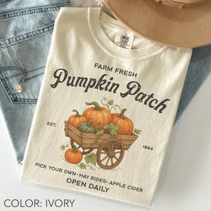 Pode incluir: Camiseta cor marfim com um desenho de abóboras numa carroça de madeira, com o texto "Farm Fresh Pumpkin Patch". Texto adicional inclui "Pick Your Own, Hay Rides, Apple Cider, Open Daily". Um chapéu bege e calças de ganga azuis estão no fundo.