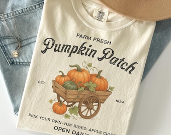 Op de afbeelding: Ivoor kleurig t-shirt met een afbeelding van pompoenen in een houten kar, met de tekst "Farm Fresh Pumpkin Patch". Extra tekst is "Pick Your Own, Hay Rides, Apple Cider, Open Daily". Een beige hoed en een blauwe spijkerbroek zijn op de achtergrond.