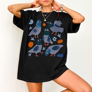 May include: Black t-shirt featuring a whimsical illustration of several cartoon pigeons in shades of gray and blue. The design includes orange and blue accent shapes. The shirt is a casual, oversized style.
