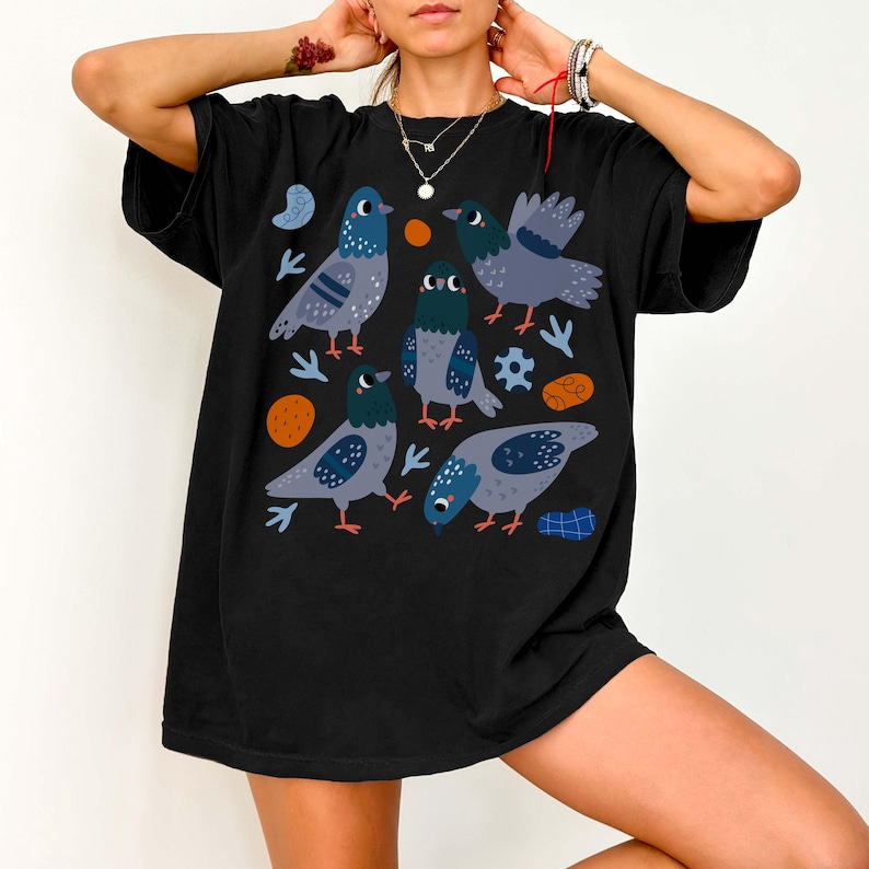 May include: Black t-shirt featuring a whimsical illustration of several cartoon pigeons in shades of gray and blue. The design includes orange and blue accent shapes. The shirt is a casual, oversized style.