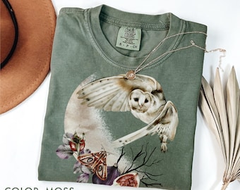 May include: A moss green t-shirt with a detailed owl graphic, moth, and floral accents. The shirt has a crew neck and short sleeves. A delicate necklace with a pendant rests on the shirt.