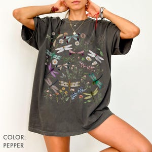 May include: Pepper-colored t-shirt featuring a colorful dragonfly and floral design. The graphic includes dragonflies in various colors, such as purple, green, and pink, amidst wildflowers and foliage. The shirt has a relaxed fit.