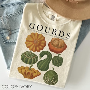 Op de afbeelding: Ivoor kleurig T-shirt met een botanische illustratie van verschillende kalebassen in oranje, groene en gele tinten. Het shirt toont de tekst "GOURDS" en "A BOTANICAL STUDY OF AUTUMN'S WHIMSICAL HARVEST".