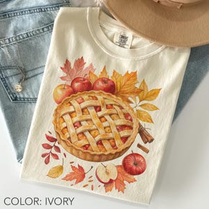 Puede incluir: Camiseta de color marfil con una ilustración de acuarela de una tarta de manzana, manzanas y hojas de otoño. La camiseta tiene cuello redondo y mangas cortas. El texto "COLOR: IVORY" está en la parte inferior.