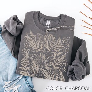 Puede incluir: Sudadera gris carb&oacute;n con dise&ntilde;o de helechos y collar plateado. El texto "COLOR: CHARCOAL" es visible. La sudadera est&aacute; colocada sobre unos vaqueros. La sudadera tiene cuello redondo.