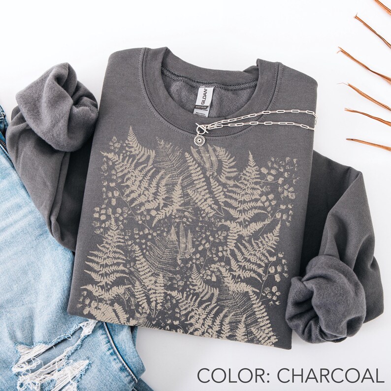 Puede incluir: Sudadera gris carb&oacute;n con dise&ntilde;o de helechos y collar plateado. El texto "COLOR: CHARCOAL" es visible. La sudadera est&aacute; colocada sobre unos vaqueros. La sudadera tiene cuello redondo.