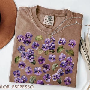 May include: Espresso-colored t-shirt adorned with a floral pattern of purple pansies and green leaves. The shirt has a crew neck and short sleeves. A silver necklace with a pendant rests on the shirt.