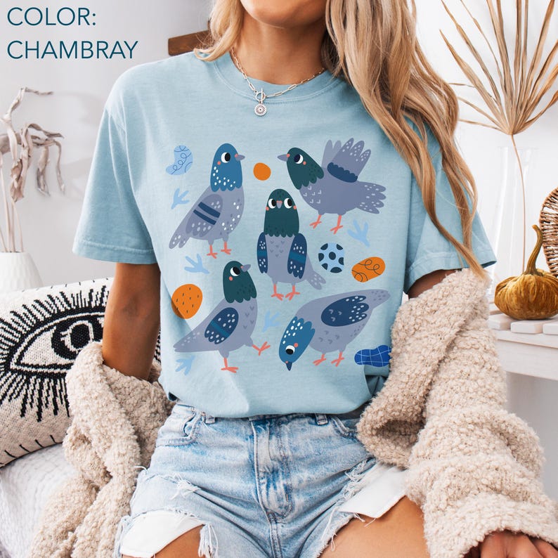May include: A light blue t-shirt with a graphic of several cartoon pigeons in shades of gray and blue. The design includes orange circles, a soccer ball, and blue leaf shapes. The shirt's color is labeled as "Chambray".