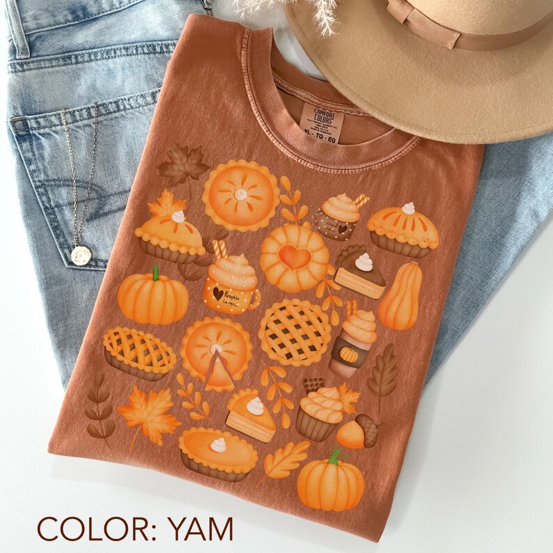 May include: A brown t-shirt with a crew neck featuring a colourful autumn-themed design. The design includes illustrations of pumpkins, pies, cupcakes, and autumn leaves. The text "COLOR: YAM" is at the bottom of the shirt.