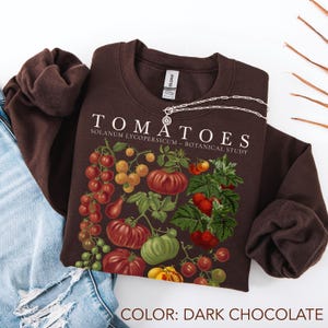 Op de afbeelding: Donkerchocoladekleurige sweatshirt met een botanische illustratie van verschillende tomaten. Het woord "TOMATOES" is in het wit gedrukt, samen met "SOLANUM LYCOPERSICUM - BOTANICAL STUDY". Een zilveren ketting ligt over de sweatshirt.