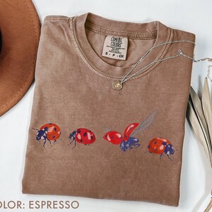 May include: Espresso-coloured t-shirt with a crew neck and a ladybug graphic. The graphic features four ladybirds in various poses, with red and black details. A silver necklace with a small pendant is draped on the shirt.