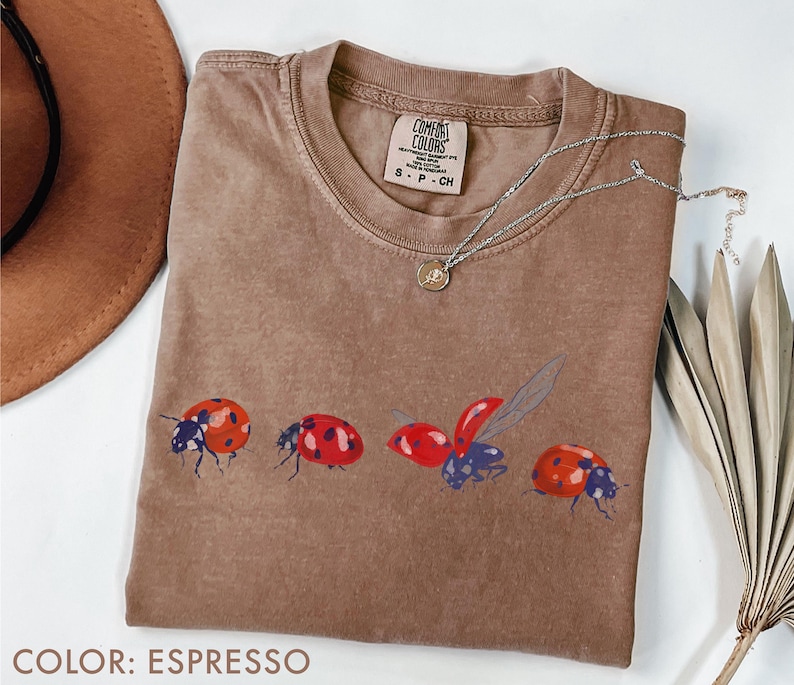May include: Espresso-coloured t-shirt with a crew neck and a ladybug graphic. The graphic features four ladybirds in various poses, with red and black details. A silver necklace with a small pendant is draped on the shirt.